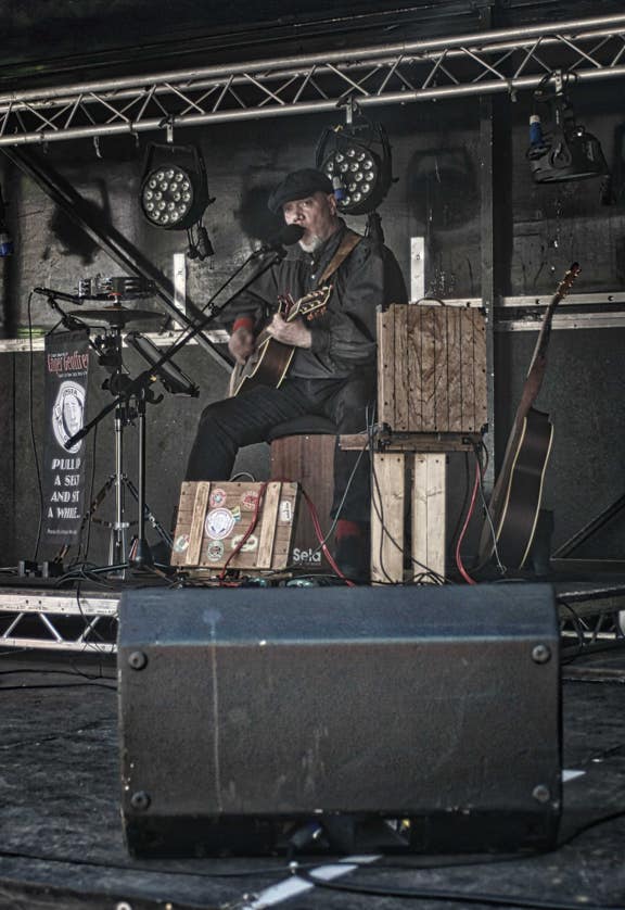 Ginger Geoffrey Sat on a cajon on stage at Jollies night club singing and playing guitar, in fornt of a backdropped of amplifier and stage lights.