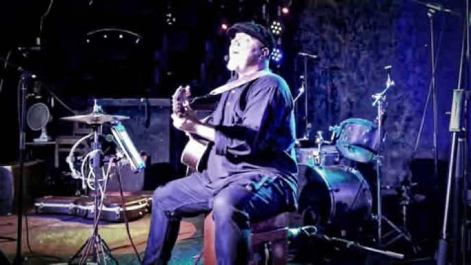 Ginger Geoffrey Sat on a cajon on stage at Jollies night club singing and playing guitar, in fornt of a backdropped of amplifier and stage lights.